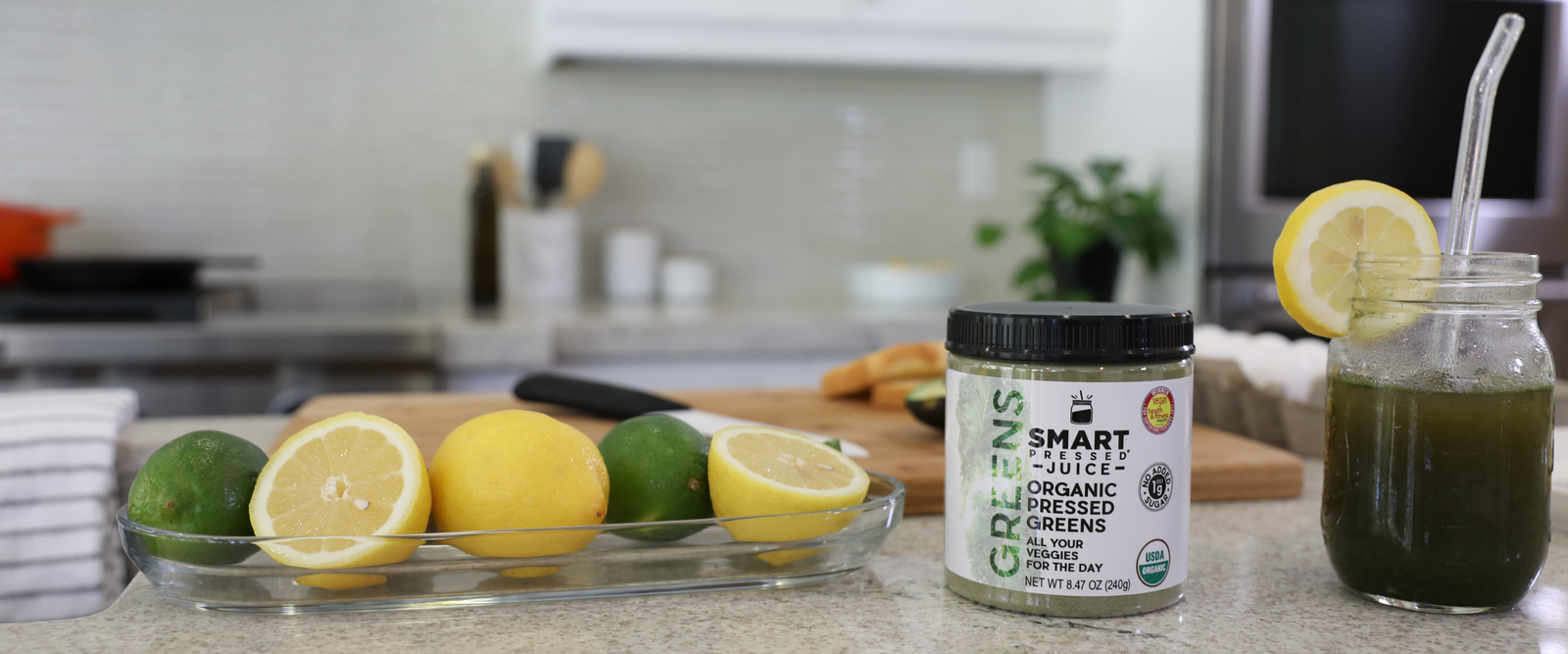 a bottle of Smart pressed organic juice and a mason jar of organic juice with a transparent straw on the side and a slice of lemon on the mouth of the mason surrounded by a plate of slices of lime and lemon