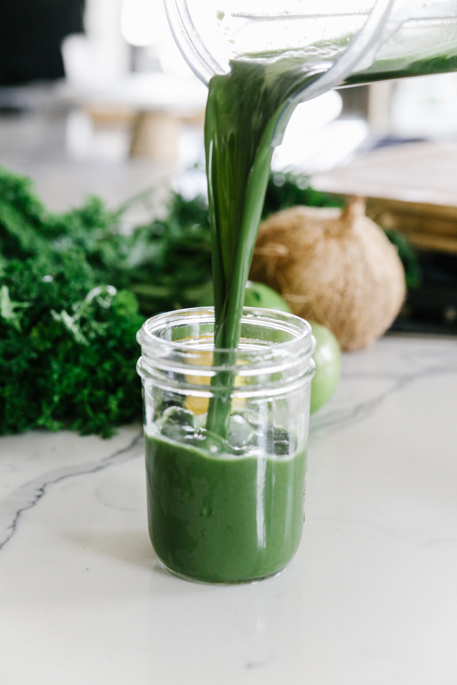 a pitcher pouring a small container of green juice and a bunch of kale,onion and green tomato on the side on top of a marble table