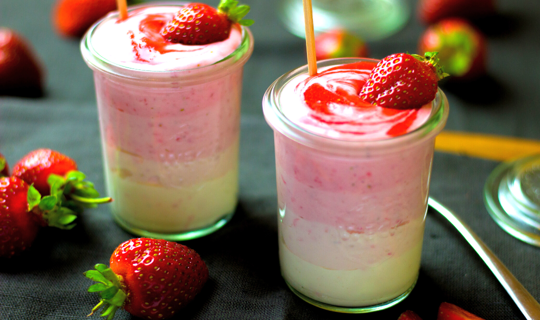 a glass of strawberry banana layered smoothie with brown paper straw held by hand surrounded by USA blocks with two strawberries in front of it and two smart labeled jar with banana and strawberries on the side with blue and red stars on the table