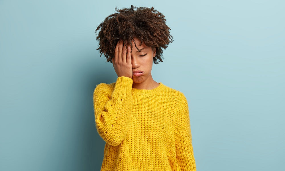 Image of a problematic-looking woman in yellow sweatshirt covering her right eye with her right hand and her left eye closed. She is standing against a blue wall.