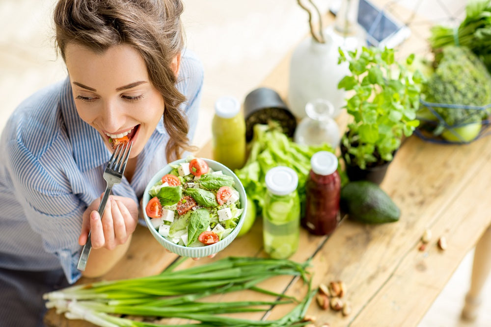 A woman enjoying her bowl of green salad - on the wooden table is some onion chives, some jars, an avocado, some herbs in a pot, and some green vegetables.