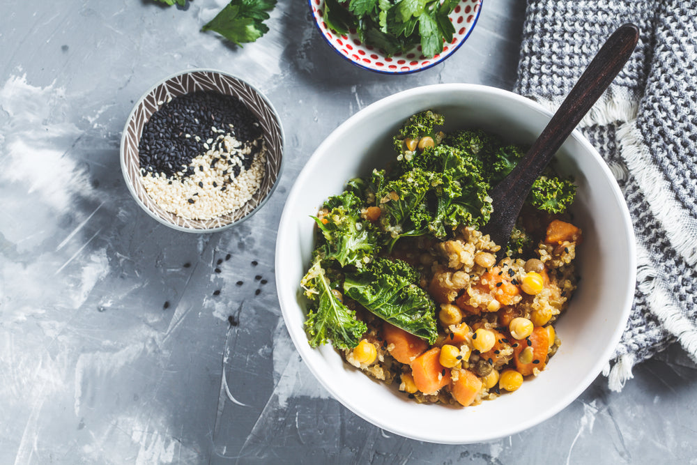 top view of bbq-spiced kale with teff and black lentils with wooden spoon surrounded by two small bowls with black and white lentils and a bunch of celery with weave black patterned cloth on top of a marble table