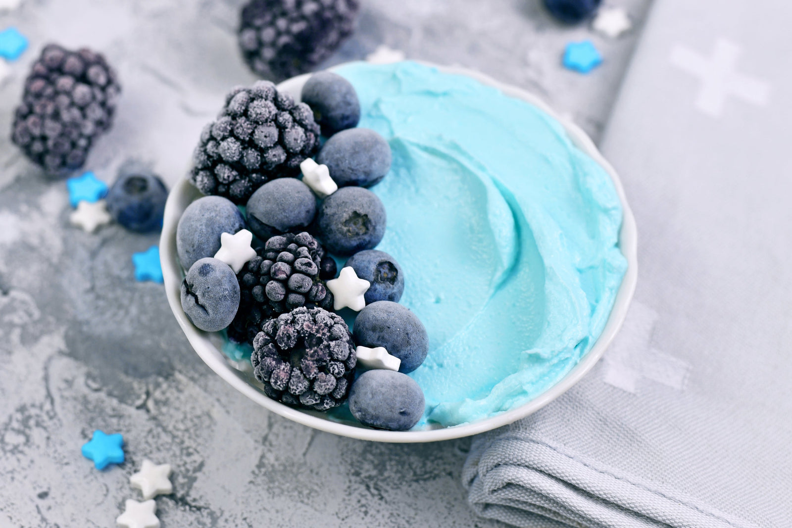a bowl of light blue-colored frozen Proteini Spirulina smoothie topped with frozen blueberries, blackberries. and star-shaped sugar flakes. On the surface are more frozen berries and star-shaped sugar flakes.