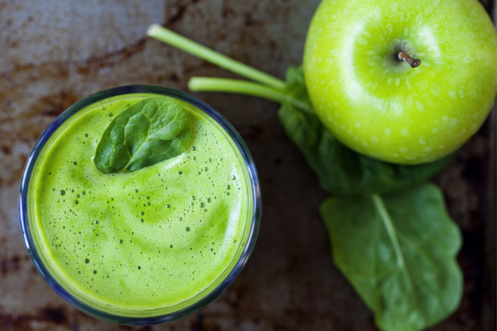 top view of a glass of apple pie pressed greens smoothie topped with a leaf of spinach with leaves of spinach topped with one green apple on the side on top of a marble table