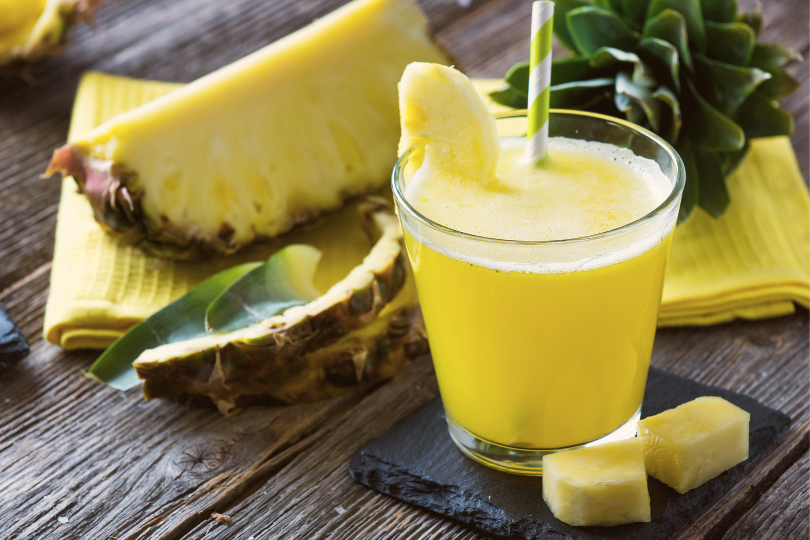 a glass of Pineapple Chia Immunity Wellness Smoothie with a straw and pineapple wedge on the rim beside 2 pineapple chunks on a black square coaster beside a cut pineapple and pineapple leaf on a yellow cloth.