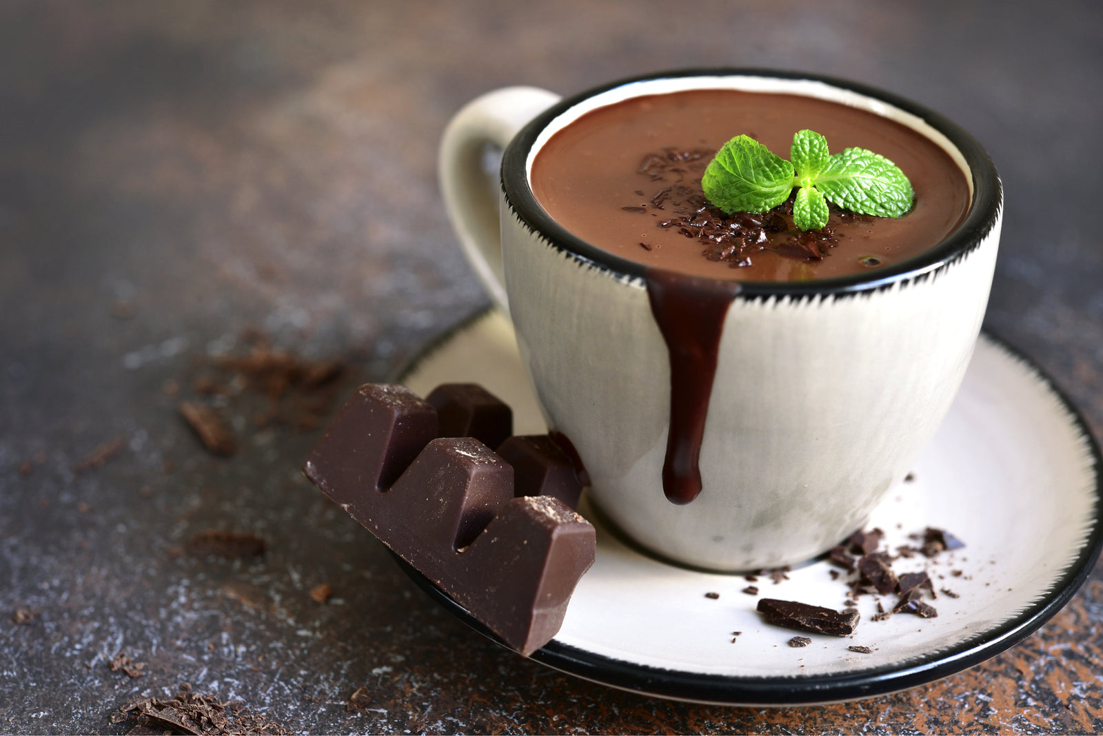 1 coffee cup of Proteini Peppermint Hot Chocolate topped with mint leaves and chocolate syrup dripping and a chunk of chocolate and chocolate crumbs on a saucer.