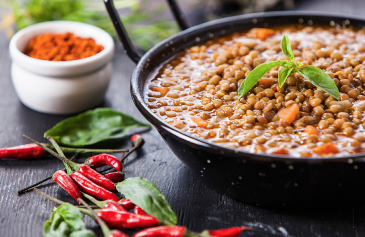 a bowl of lentil soup garnished with green herb beside bird-eye chilies and a bowl of sweet paprika on the wooden surface.