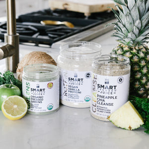 One 240 grams open jar of Organic Pressed Greens, one 500 grams open jar of Vegan Vanilla Proteini, and one 375 grams open jar of Pineapple Cleanse side by side surrounded by a slice of pineapple, parsley, whole pineapple, 1 coconut, 1 apple, and half an orange in a kitchen setting.