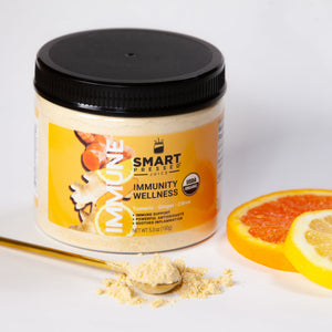 1 jar of 150 grams Immunity Wellness, in front is a stirring spoon full of light brown powder, and 2 slices of oranges on the side on a white surface.