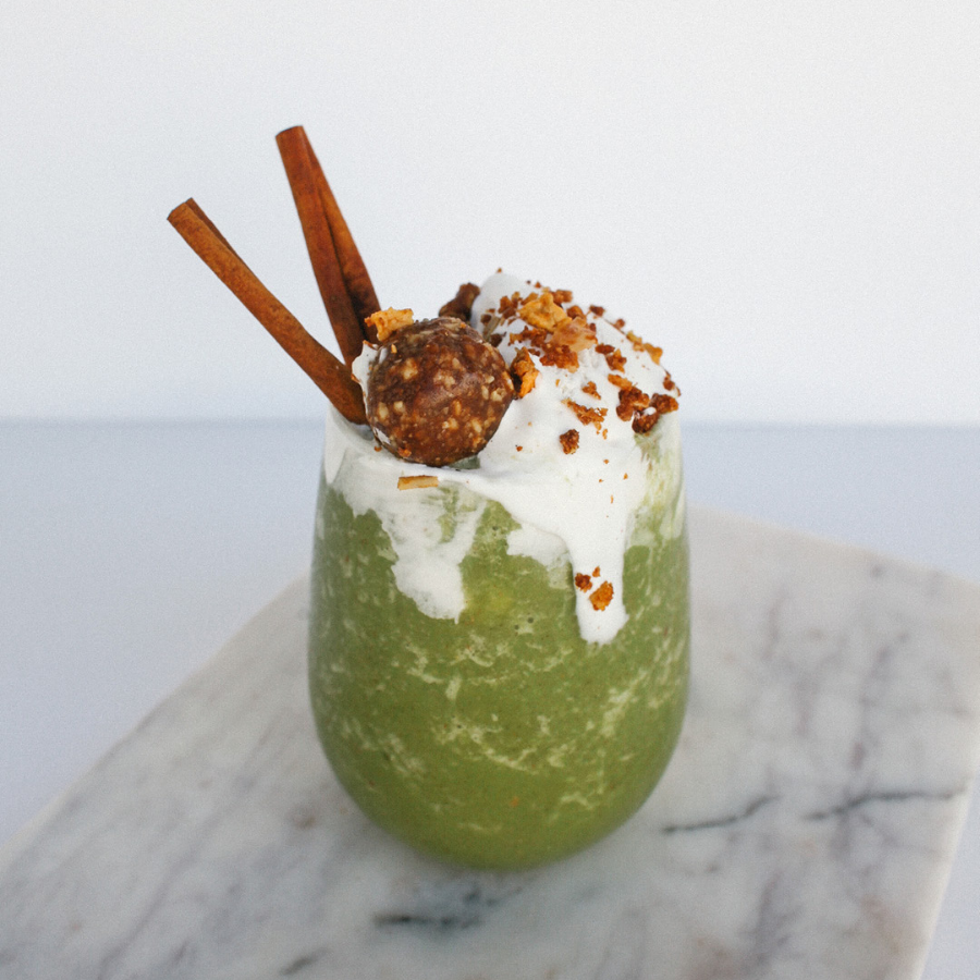 a glass of apple pie smoothie topped with whip cream, ball-shaped choco nuts, two vanilla sticks, and apple cinnamon granola on top of a marble table