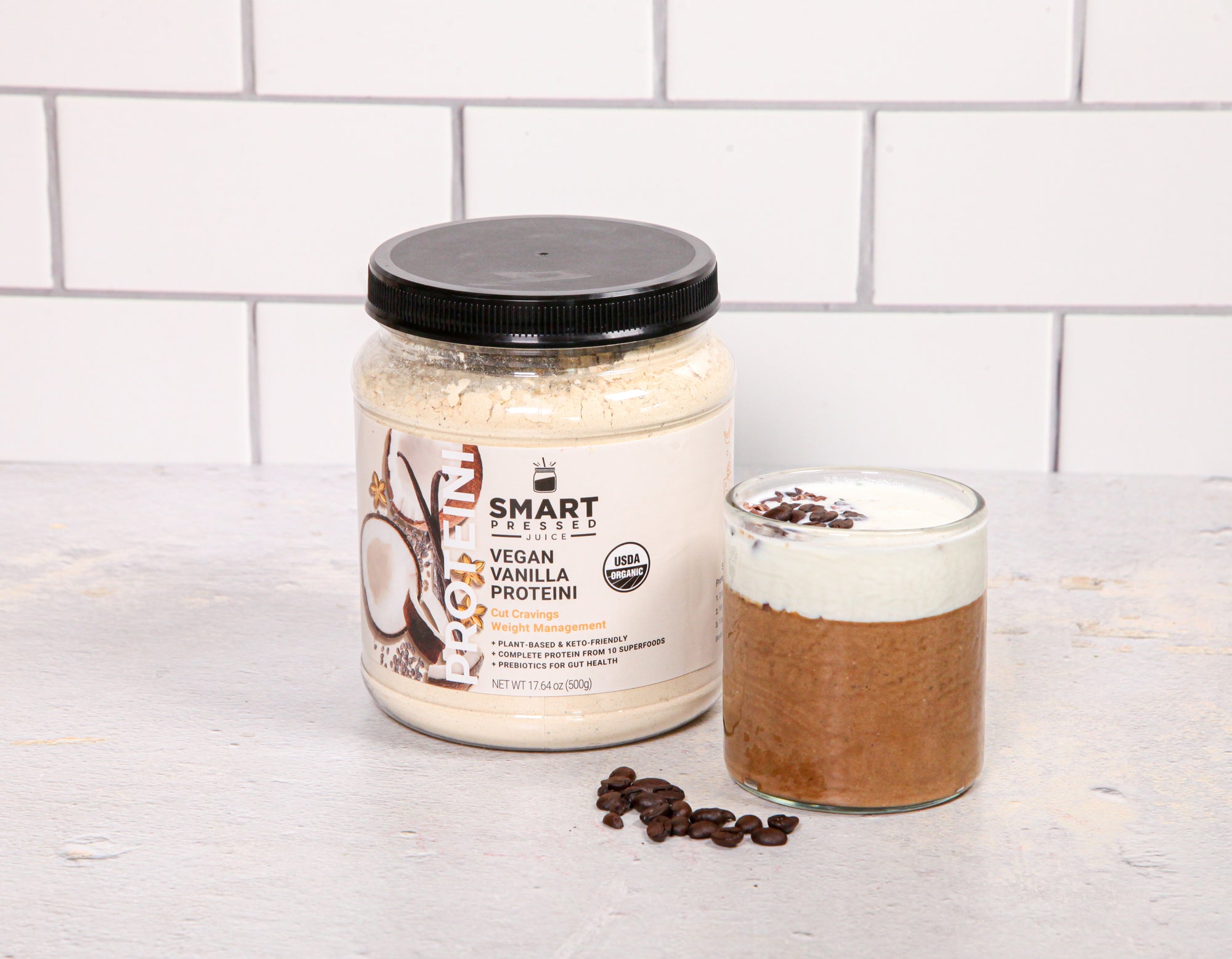 a glass of coffee proteini with a jar of smart labeled vegan vanilla proteini with roasted coffee beans on the side on top of a white painted table