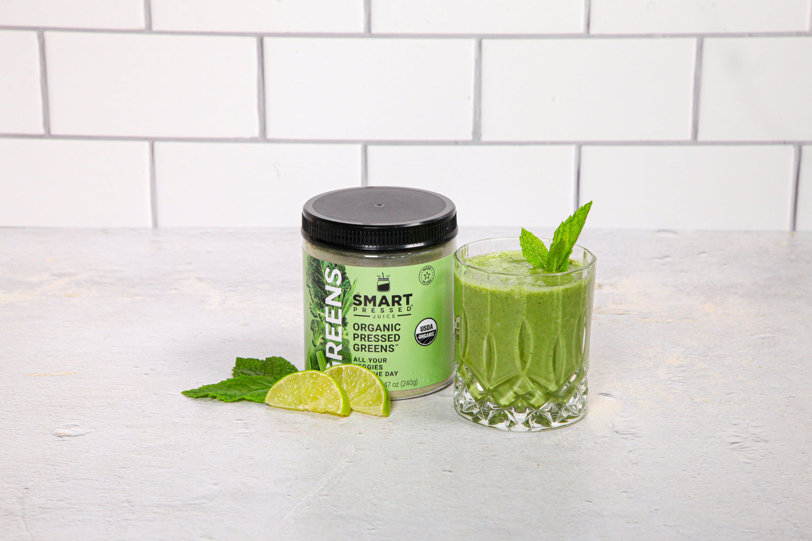 front view of a glass of Signature Greens Smoothie topped with mint and a jar of smart pressed organic pressed green with two slices of lime on top of mint leaves on top of a white painted table