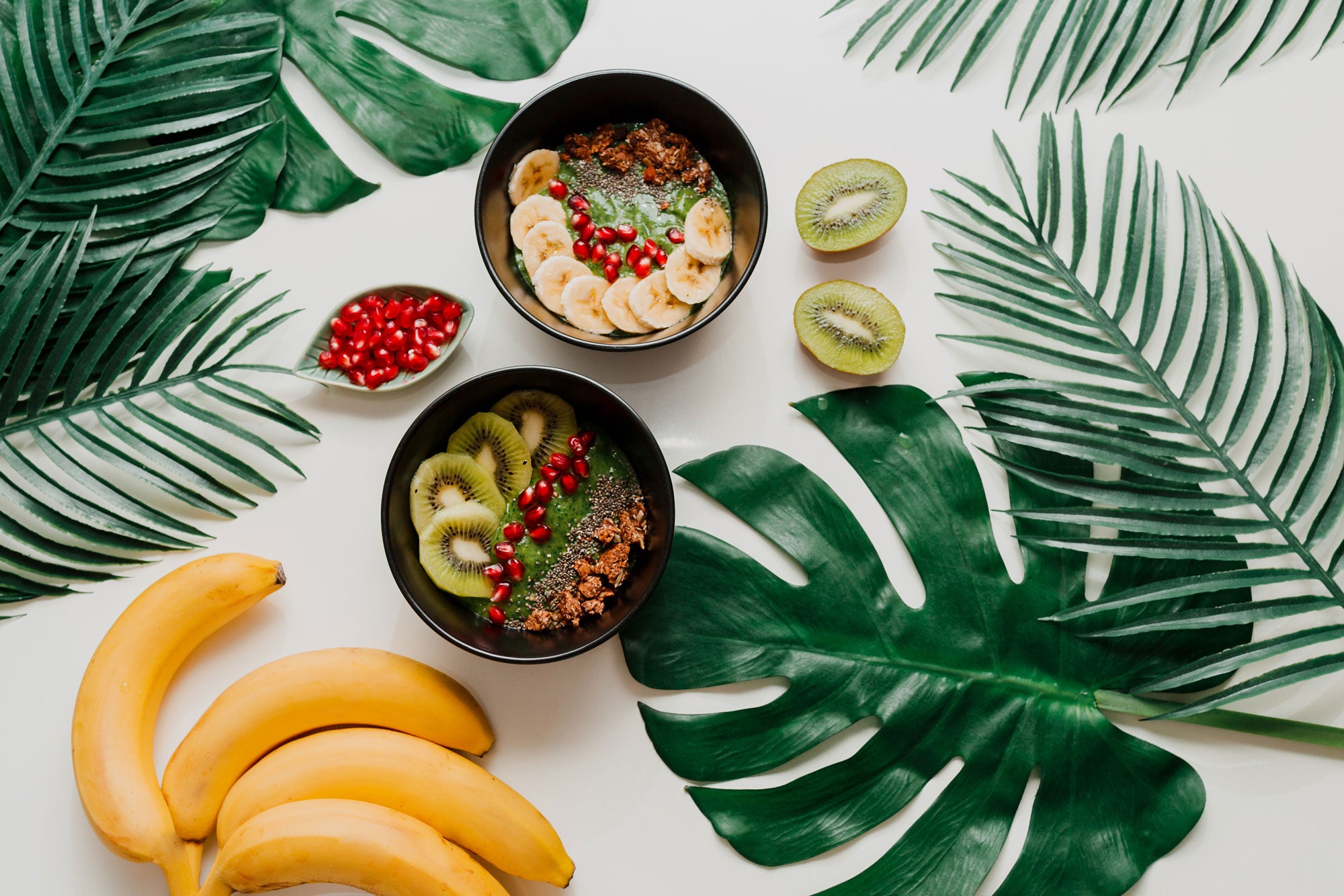 Image of 2 bowls of fruits and oats surrounded by green leaves and a pile of banana and 2 slices of kiwi.