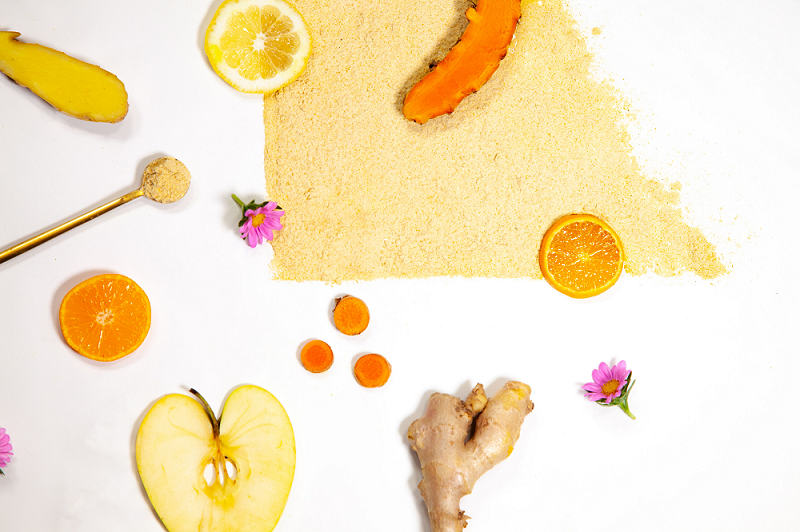 A left triangular shaped light brown powder on a white surface with a slice of lemon, orange, and turmeric on top. On the white surface is a slice of apple, ginger and carrot, a stirring spoon full of light brown powder, and 2 purple flower.