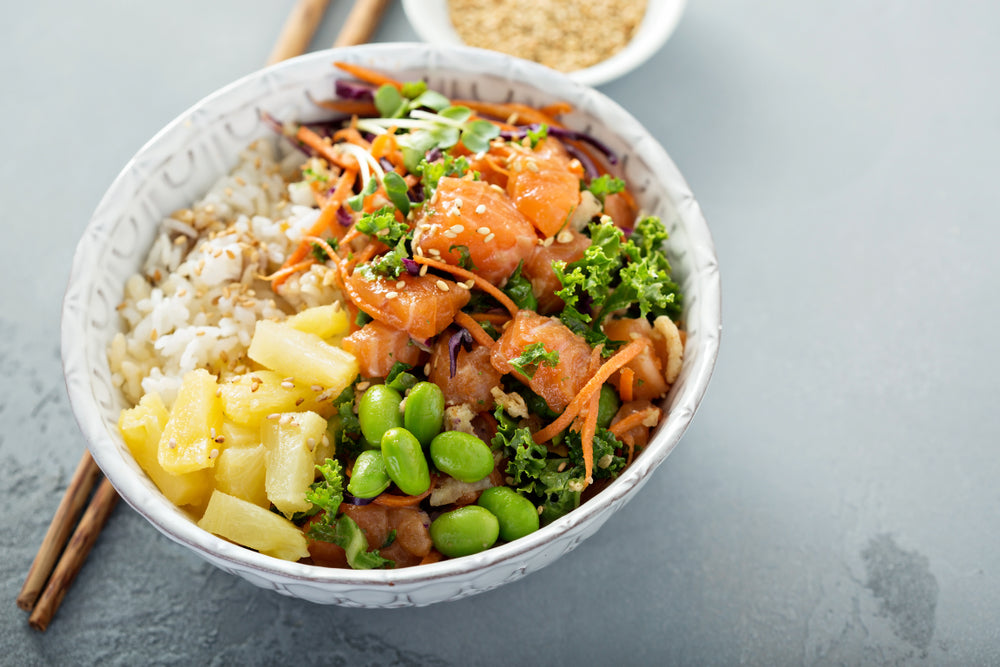 top view of a ceramic bowl with pineapple-ginger rice bowl with chopsticks on the side a  small bowl of sesame seeds on top of a blue-painted table 