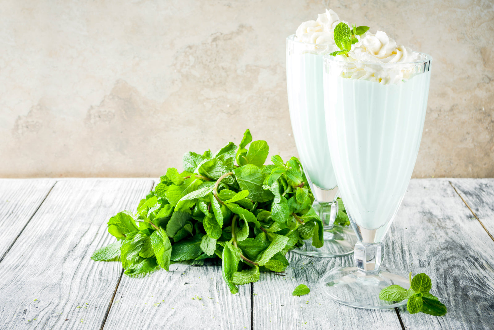 side view of two glasses of vegan shamrock shake topped with whip cream and mint surrounded by a bunch of mint leaves on top of a wooden table 