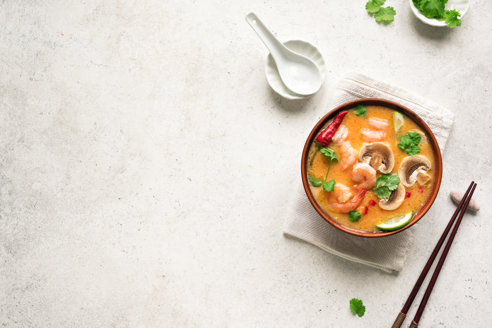 top view of a bowl of tom ka soup topped with slices of mushroom, shrimps, slices of lemon, cilantro, and chili on top of a white folded table cloth with chopsticks and soup spoon with a saucer on the side and a white bowl with cilantro