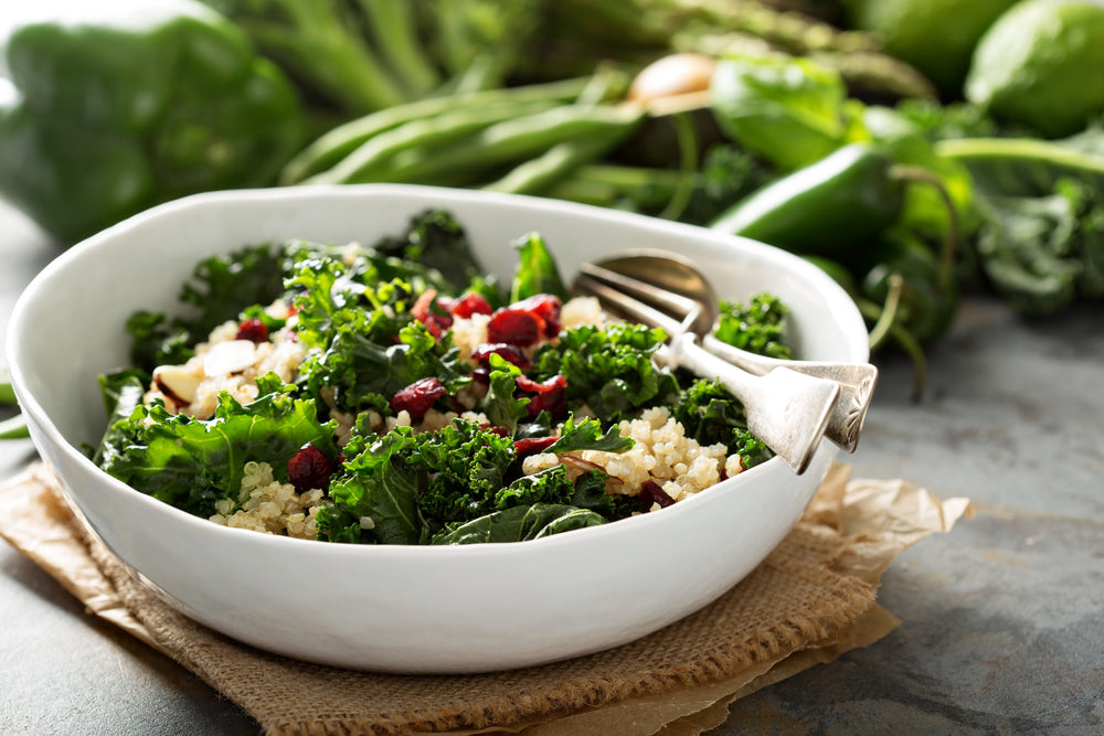 side view of a white bowl with kale Waldorf salad with spoon and fork on top of a weaved cloth with  paper on the bottom with green fresh vegetables on the side of the bowl