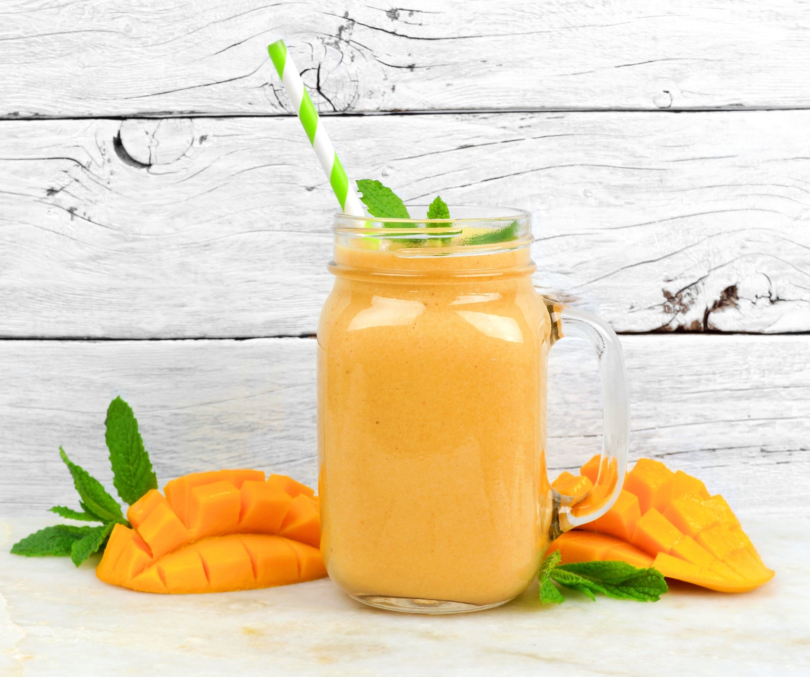 1 majon jar glass of Michelle's Tropical Immunity Smoothie garnished with green leaves beside 1 sliced ripe mango on both sides. 