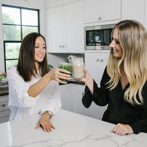 Two beautiful and healthy-looking ladies, each holding a jar of smoothies making a toast.