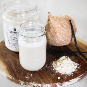 One 500 grams open jar of Vegan Vanilla Proteini beside a coconut with 2 sticks of vanilla standing against it in the back row. In the front row is a jar of white liquid and light brown powder on a circular wood coaster. Set against a white background.