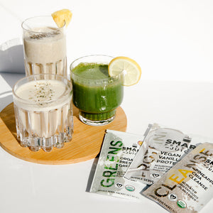 1 tall glass of white smoothie with chia seed, a straw, and a slice of pineapple, small glass of white smoothie with green garnish, and a small glass of green smoothie with a slice of lemon. The 3 glasses are all placed in one wooden coaster. Beside is 3 single serving sachets  of Organic Pressed Greens, Vegan Vanilla Proteini, and Pineapple Chia Cleanse on a white surface.