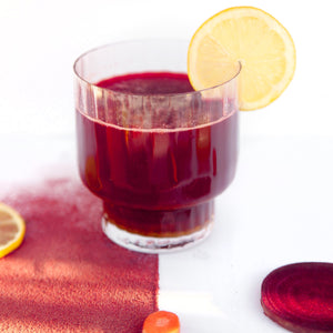 A glass of pink juice with a slice of lemon and a slice of beet root, a slice of lemon, and a slice or turmeric on the half-white-half-dark pink surface.