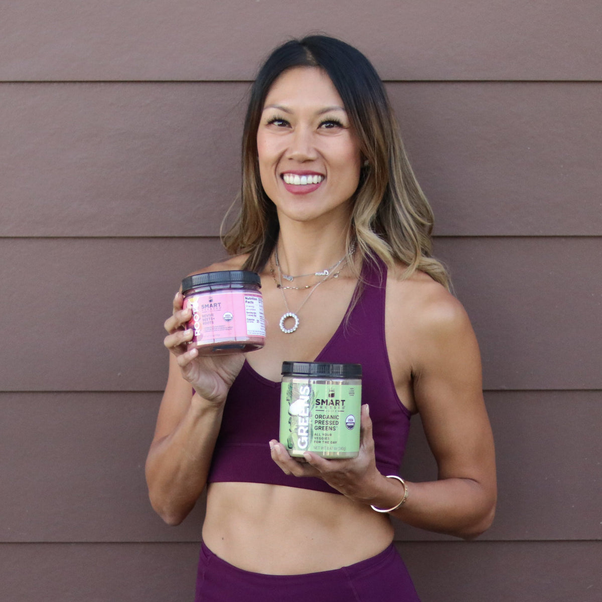 A beautiful and healthy-looking woman holding a jar of Revive Beet+Roots on the right and a jar of Organic Pressed Greens on the left. She is standing against a brown wall.