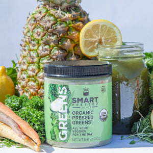 1 jar of 240 grams Organic Pressed Greens beside a jar of green smoothie with a slice of orange, beside is a pineapple fruit, broccoli, carrots, and lemon side by side against a light blue background. 