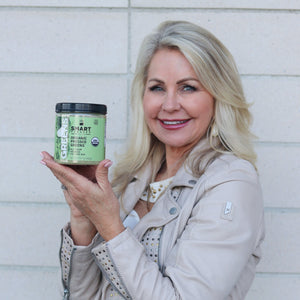 A beautiful and healthy-looking elderly woman holding a jar of 240 grams Organic Pressed Greens standing against a brick wall.